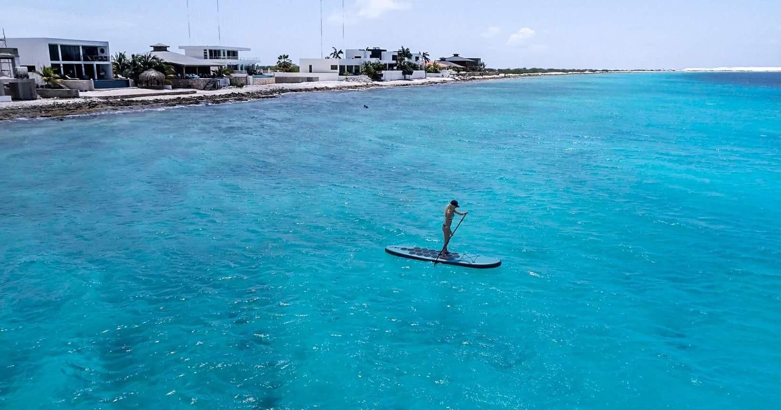 Luxury Bonaire villa guest paddleboarding on the calm turquoise Caribbean Sea directly in front of modern oceanfront properties.